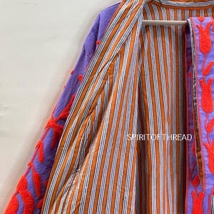 May include: A colorful, open-front jacket with a striped orange and white interior. The exterior features purple fabric with red floral embroidery. The jacket is hanging on a wooden hanger.