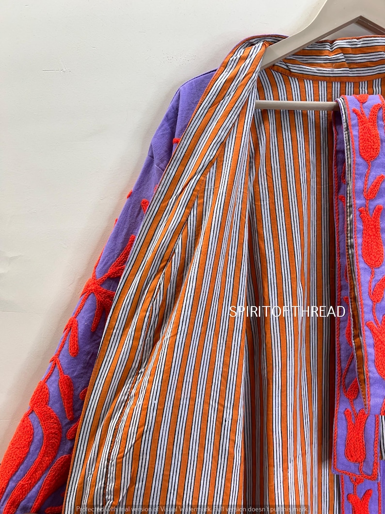 May include: A colorful, open-front jacket with a striped orange and white interior. The exterior features purple fabric with red floral embroidery. The jacket is hanging on a wooden hanger.