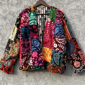 May include: A vibrant patchwork jacket with floral embroidery. The jacket is made up of various fabric squares in shades of red, pink, blue, green, and yellow. It hangs on a wooden hanger.