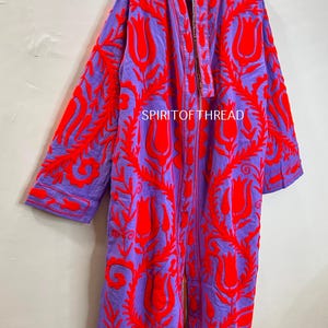 May include: A long, open-front robe in a vibrant purple hue, adorned with bold red floral embroidery. The robe features long sleeves and a detailed pattern, with the text "SPIRITOFTHREAD" visible. This garment is a striking piece of apparel.
