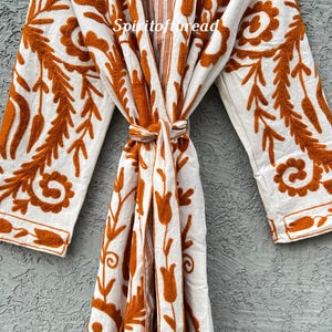 Puede incluir: Bata blanca con intrincados bordados florales en naranja. La bata presenta mangas largas y un cierre de lazo en la cintura. El texto "Spiritofthread" es visible. La bata est&aacute; hecha de una tela suave y fluida.