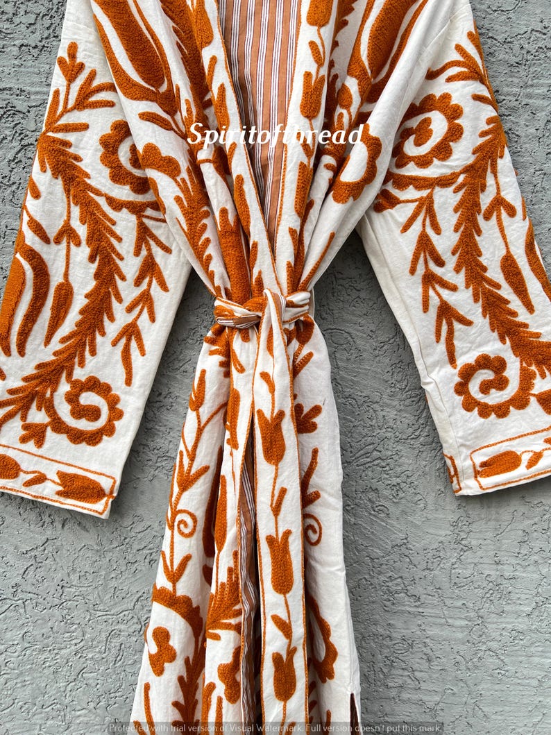 Puede incluir: Bata blanca con intrincados bordados florales en naranja. La bata presenta mangas largas y un cierre de lazo en la cintura. El texto "Spiritofthread" es visible. La bata est&aacute; hecha de una tela suave y fluida.