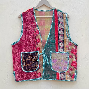 Könnte beinhalten: Eine Patchwork-Weste mit floralen und geometrischen Mustern in Rot-, Grün- und Beigetönen. Die Weste hat zwei Vordertaschen und ist mit einer türkis-weiß gestreiften Bordüre versehen. Sie hängt an einem Holzkleiderbügel.