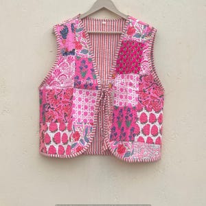 May include: A pink patchwork vest with various floral and geometric patterns. The vest has a striped interior and is fastened with wooden buttons. The vest is hanging on a wooden hanger.