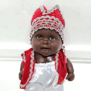 Pode incluir: Uma pequena boneca vestindo uma coroa vermelha e prateada, um colete vermelho e prateado e calças brancas. A boneca tem pele escura e uma expressão sorridente.
