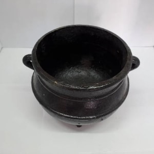 Może przedstawiać: Czarny kocioł żeliwny z dwoma uchwytami i trzema nogami. Kocioł jest pusty i ma gładką, błyszczącą powierzchnię.