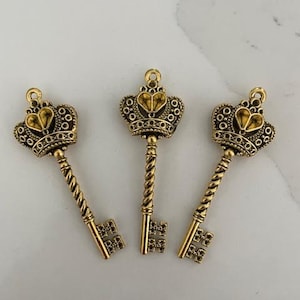 Puede incluir: Tres dijes de llave de metal en tono dorado, cada uno con un arco en forma de corona con un detalle central en forma de corazón. Los vástagos de las llaves tienen una textura de cuerda trenzada y los paletones son rectangulares con un patrón de rejilla.