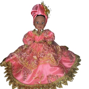 Puede incluir: Una muñeca de piel oscura y ojos marrones que lleva un pañuelo de satén rosa y un vestido de satén rosa a juego. El vestido presenta mangas abullonadas, una falda ancha con bordados florales dorados y un borde de encaje dorado festoneado.