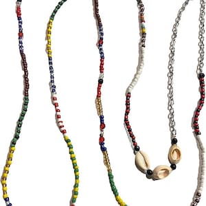 Puede incluir: Un collar largo que presenta una hebra de cuentas de vidrio multicolores en rojo, azul, verde, amarillo y blanco, junto a una segunda hebra que combina una cadena de metal plateado con cuentas negras y tres conchas de cauri naturales.