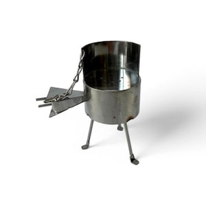 Puede incluir: Un pequeño hornillo de camping cilíndrico de metal con una base perforada, tres patas finas de apoyo y una bandeja de combustible triangular montada en el lateral y sujeta por una cadena metálica. La estructura es de acero inoxidable pulido.