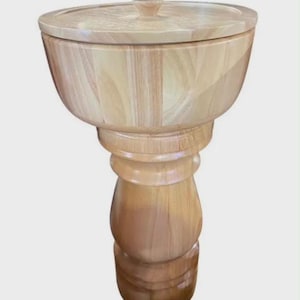 May include: A light-colored wooden pedestal bowl with a matching circular lid featuring a small central knob. The bowl has a wide, flared top section that tapers down into a thick, rounded, and tiered support base with a smooth, polished finish.