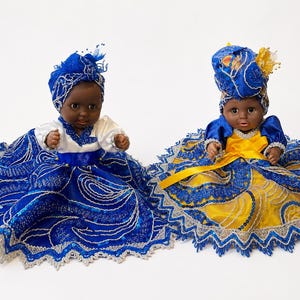 May include: Two dolls with dark skin tones wearing matching blue and white headwraps. One doll wears a blue and white dress with sequin details, while the other wears a blue and yellow dress with a yellow sash and a silver lace trim border.