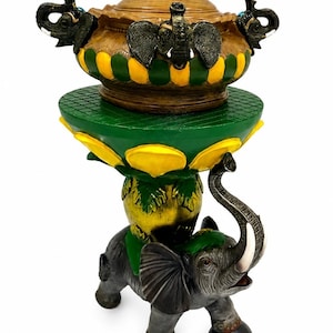 Puede incluir: Un soporte decorativo con una figura de elefante gris con colmillos blancos que equilibra una plataforma verde y amarilla sobre su espalda. Sobre la plataforma hay un cuenco con tapa de rayas marrones, verdes y amarillas y tres elefantes.