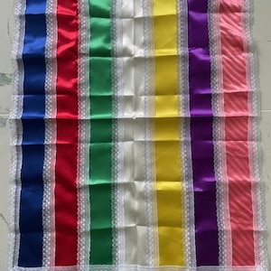 Puede incluir: Una pieza de tela rectangular con rayas verticales de satén en azul, rojo, verde, blanco, amarillo, morado y rosa. Cada raya está separada por un fino borde de encaje blanco y todo el perímetro está ribeteado con encaje blanco a juego.