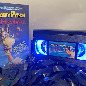Może przedstawiać: Czarna lampa w kształcie kasety VHS z niebieskim światłem w środku. Przód lampy przedstawia obraz filmu "Monty Python i Święty Graal" z tekstem tytułu. Lampa stoi na łóżku z czarnej taśmy VHS.