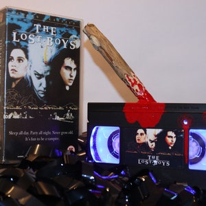 Puede incluir: Una cinta VHS negra de la película "The Lost Boys" con una estaca de madera cubierta de pintura roja. La cinta tiene una luz azul en el interior y está rodeada de tiras de película negras. La portada muestra el título y los actores de la película.