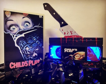 May include: A black VHS tape with a bloody knife sticking out of it. The tape has a blue light shining from inside and a Child's Play movie poster on the front. The tape is surrounded by black VHS tapes.