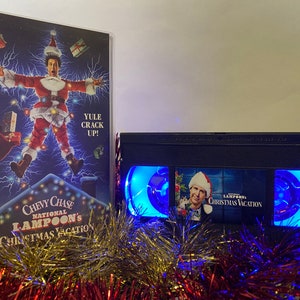 May include: A black VHS tape with a blue light shining from the windows. The tape has a picture of Chevy Chase as Clark Griswold from the movie National Lampoon's Christmas Vacation. The text on the tape reads "National Lampoon's Christmas Vacation".