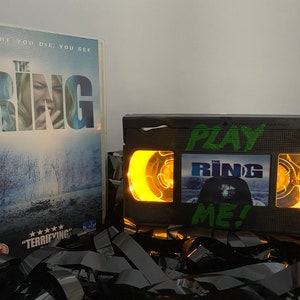 Peut inclure: Une cassette VHS noire avec une lumière jaune qui brille à travers les fenêtres. La cassette porte l'inscription "Play The Ring Me !". La cassette est posée sur une pile de cassettes VHS noires.