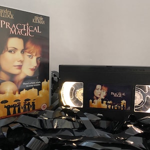 Puede incluir: Una cinta VHS de la película "Practical Magic" protagonizada por Sandra Bullock y Nicole Kidman. La cinta está en una caja negra con una etiqueta blanca. La etiqueta presenta una foto de las dos actrices y el título de la película. La cinta está rodeada de cintas VHS negras.