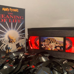 May include: A black VHS tape with a red label that says "Monty Python's The Meaning of Life". The tape is sitting on a pile of black VHS tapes. The cover art for the tape features a group of people standing on a hill.