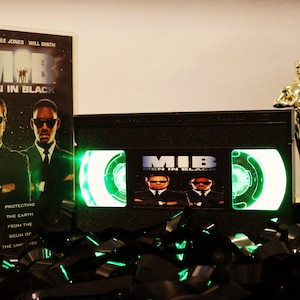 May include: A VHS tape of the movie Men in Black with a green light shining through the window. The tape is surrounded by black and green film. The movie cover is also visible with the actors Tommy Lee Jones and Will Smith on it. The text on the cover reads "Men in Black".