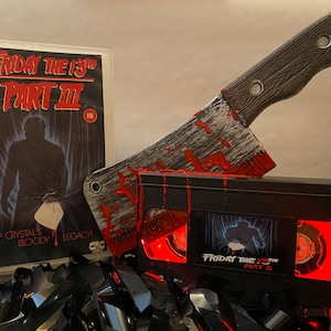 以下が含まれることがあります： 赤い光を放つ黒いVHSテープ。ラベルには「13日の金曜日 パート3」の文字。血まみれの肉屋の包丁がテープの上に置かれている。テープは黒いVHSテープに囲まれている