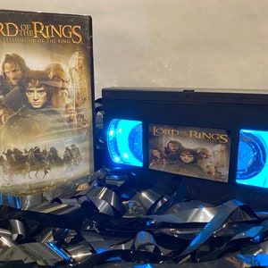 May include: A black VHS tape with a blue light shining through the windows. The tape has a label that says "The Lord of the Rings: The Fellowship of the Ring".
