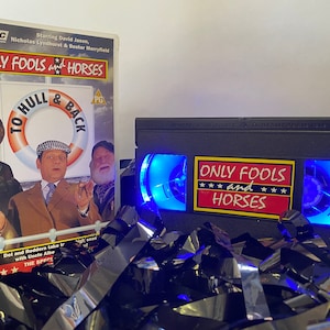May include: A VHS tape of the British sitcom "Only Fools and Horses" with a blue light shining through the clear plastic case. The tape is sitting on a bed of black and silver streamers.