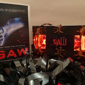 May include: A black VHS tape with red blood splatter and barbed wire wrapped around it. The tape has a red light shining through the window and the word "SAW" is printed on the front. The tape is sitting on a pile of black VHS tape.