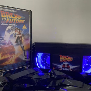 Può includere: Un nastro VHS nero con una luce blu che brilla attraverso le finestre di plastica trasparente. Il nastro ha un adesivo sul davanti che dice "Ritorno al futuro" con un'immagine della DeLorean. Il nastro è su una superficie nera con una custodia DVD per il film "Ritorno al futuro" sullo sfondo.