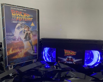 May include: A black VHS tape with a blue light shining through the clear plastic windows. The tape has a sticker on the front that says "Back to the Future" with a picture of the DeLorean time machine. The tape is sitting on a black surface with a DVD case for the movie "Back to the Future" in the background.