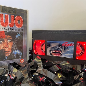 Puede incluir: Una cinta VHS con una luz roja brillando a través de la ventana. La cinta está etiquetada como "Cujo" y presenta un perro gruñendo con una expresión amenazante. La cinta está sentada sobre una pila de cintas VHS negras y plateadas.