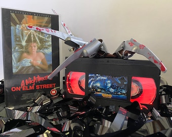 May include: A VHS tape of the film "A Nightmare on Elm Street" with a Freddy Krueger glove prop on top. The tape is surrounded by black and silver VHS tape.