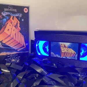 Może przedstawiać: Czarna kaseta VHS z niebieskim światłem świecącym przez przezroczyste plastikowe okienko. Kaseta ma etykietę z tytułem "Życie Briana" i obraz tytułowej karty filmu. Kaseta leży na stosie czarnych kaset VHS.