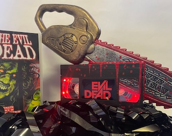 May include: A black VHS tape with a red and black splatter paint design. The tape has the words "Evil Dead" printed on it. A red and gold chainsaw prop is on top of the tape. A comic book with the words "The Evil Dead" is on the left side of the image.