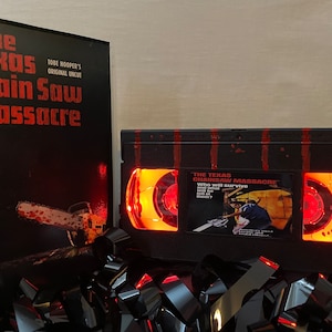 Puede incluir: Una réplica de cinta VHS negra de la película de terror de 1974 "The Texas Chainsaw Massacre". La cinta está iluminada desde adentro con un brillo rojo, y el título "The Texas Chainsaw Massacre" y el texto "Who will survive" son visibles en la cinta. La cinta está sentada sobre una pila de cintas VHS negras.