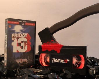 May include: A black VHS tape with a red blood splatter design and the text "Friday the 13th" on the front. A black axe with a brown handle is stuck into the tape. The tape is surrounded by black film reels.