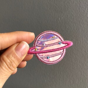 May include: Pink iridescent embroidered iron-on patch of a planet with rings. The planet has a holographic finish and is surrounded by a pink ring.