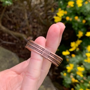 Op de afbeelding: Een koperen draadring met een gevlochten ontwerp. De ring wordt aan een vinger gedragen.