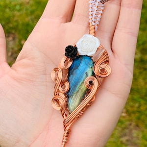 May include: Copper wire-wrapped pendant with a blue and green labradorite stone. The pendant features white and black rose accents and a copper chain. The wirework is intricate, with a woven top section and decorative swirls.