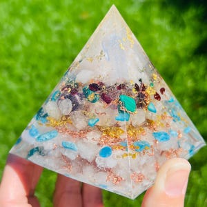 K&ouml;nnte beinhalten: Eine klare pyramidenf&ouml;rmige Kristall mit eingebetteten Kristallen in verschiedenen Farben, darunter Blau, Gr&uuml;n und Rot. Die Pyramide ist mit goldenen und kupfernen Flocken gef&uuml;llt.