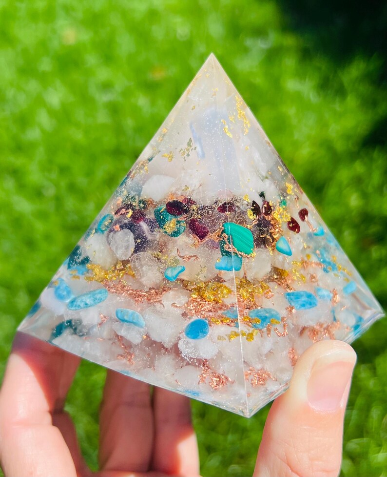K&ouml;nnte beinhalten: Eine klare pyramidenf&ouml;rmige Kristall mit eingebetteten Kristallen in verschiedenen Farben, darunter Blau, Gr&uuml;n und Rot. Die Pyramide ist mit goldenen und kupfernen Flocken gef&uuml;llt.