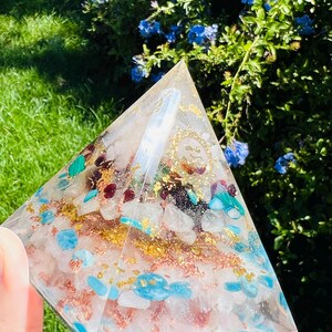 K&ouml;nnte beinhalten: Eine klare pyramidenf&ouml;rmige Kristall mit goldenen Flocken und verschiedenen Kristallen, die darin eingebettet sind, darunter T&uuml;rkis, Rosenquarz und Malachit.