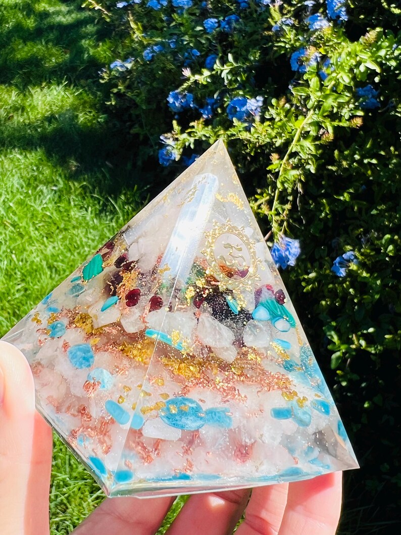 K&ouml;nnte beinhalten: Eine klare pyramidenf&ouml;rmige Kristall mit goldenen Flocken und verschiedenen Kristallen, die darin eingebettet sind, darunter T&uuml;rkis, Rosenquarz und Malachit.