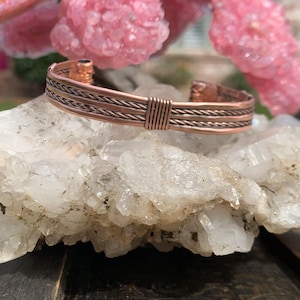 Op de afbeelding: Een koperen armband met een gevlochten ontwerp. De armband ligt op een bed van witte kristallen.