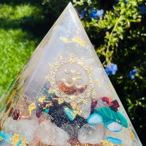 K&ouml;nnte beinhalten: Eine klare Kristallpyramide mit goldenen Akzenten und eingebetteten Kristallen. Die Pyramide hat ein goldenes Om-Symbol in der Mitte.