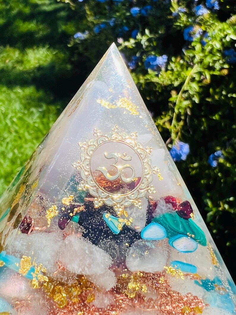 K&ouml;nnte beinhalten: Eine klare Kristallpyramide mit goldenen Akzenten und eingebetteten Kristallen. Die Pyramide hat ein goldenes Om-Symbol in der Mitte.
