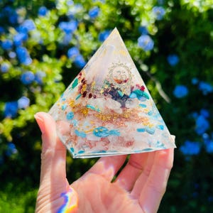 K&ouml;nnte beinhalten: Eine klare Kristallpyramide mit verschiedenen Kristallen, die darin eingebettet sind, darunter Rosenquarz, Amethyst und T&uuml;rkis. Die Pyramide liegt auf einer Hand, und der Hintergrund ist ein verschwommenes Bild von gr&uuml;nem Laub.