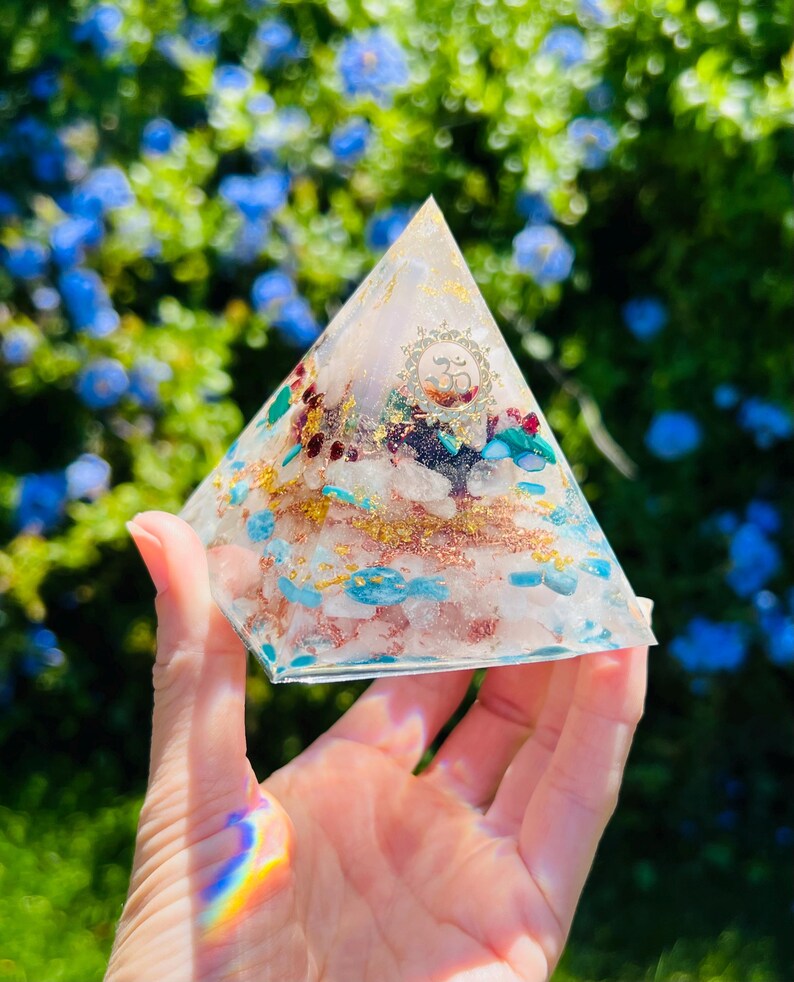 K&ouml;nnte beinhalten: Eine klare Kristallpyramide mit verschiedenen Kristallen, die darin eingebettet sind, darunter Rosenquarz, Amethyst und T&uuml;rkis. Die Pyramide liegt auf einer Hand, und der Hintergrund ist ein verschwommenes Bild von gr&uuml;nem Laub.